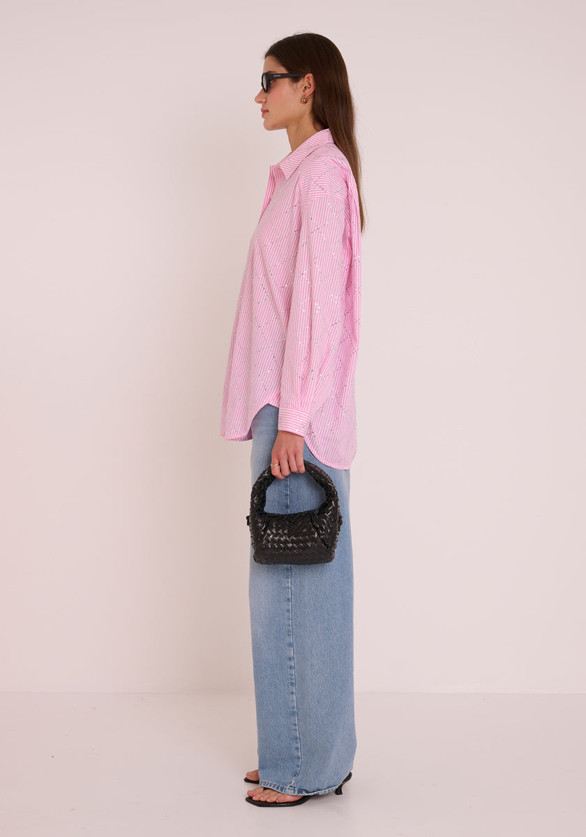 Eine Frau im Profil vor einem schlichten hellen Hintergrund trägt eine rosa gestreifte Princess Goes Hollywood-Bluse, hellblaue Jeans mit weitem Bein, schwarze Sandalen mit offenen Zehen und Sonnenbrille und hält eine kleine schwarze gewebte Handtasche in ihrer linken Hand.