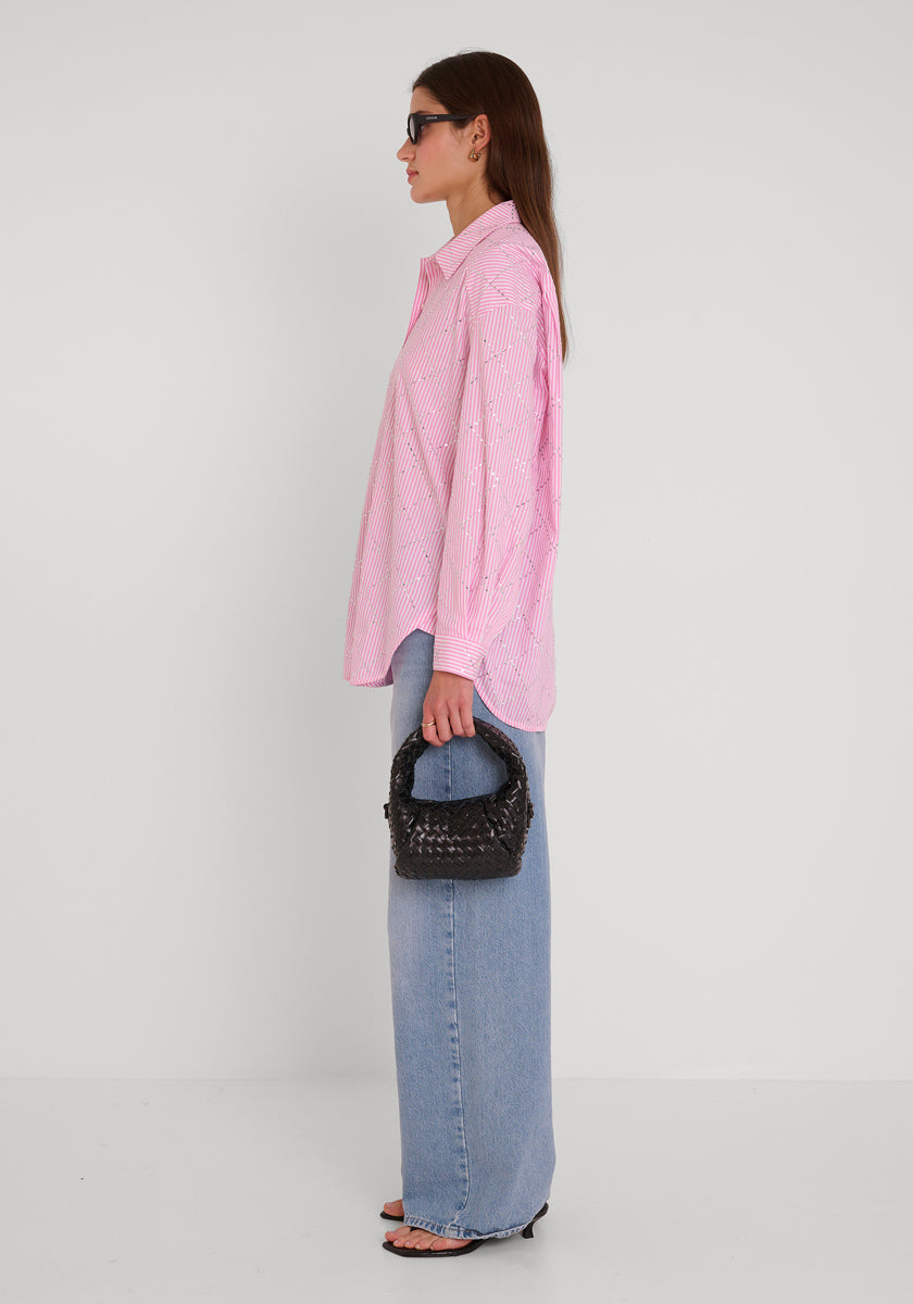 Eine Frau im Profil posiert vor einer weißen Wand. Sie trägt ein gestreiftes Princess Goes Hollywood-Shirt, hellblaue Jeans mit weitem Bein, schwarze offene Absätze, eine dunkle Sonnenbrille und hält eine kleine schwarze gewebte Handtasche in der linken Hand.