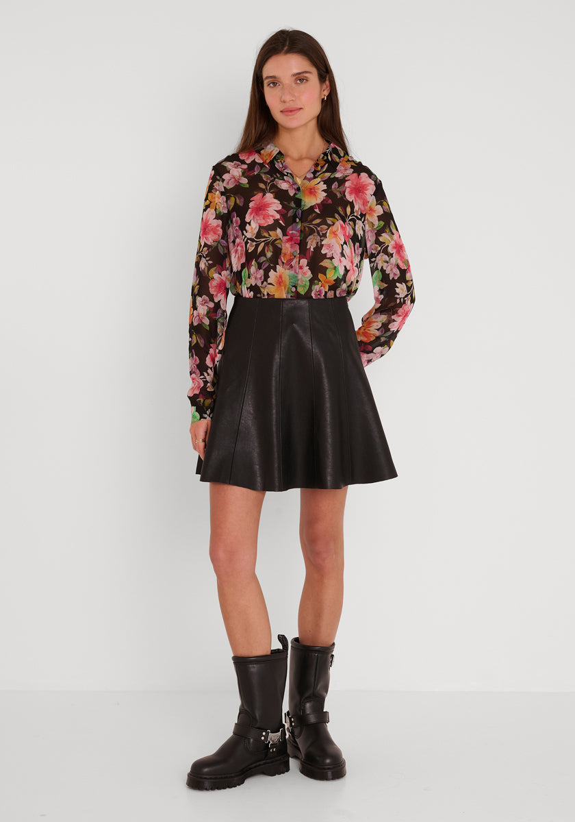 Eine Frau mit langen braunen Haaren und neutralem Gesichtsausdruck steht vor einer weißen Wand und trägt eine langärmelige Bluse mit Blumendruck von Princess Goes Hollywood, dazu einen schwarzen ausgestellten Lederrock und schwarze kniehohe Biker-Stiefel mit Riemen und Metalldetails.