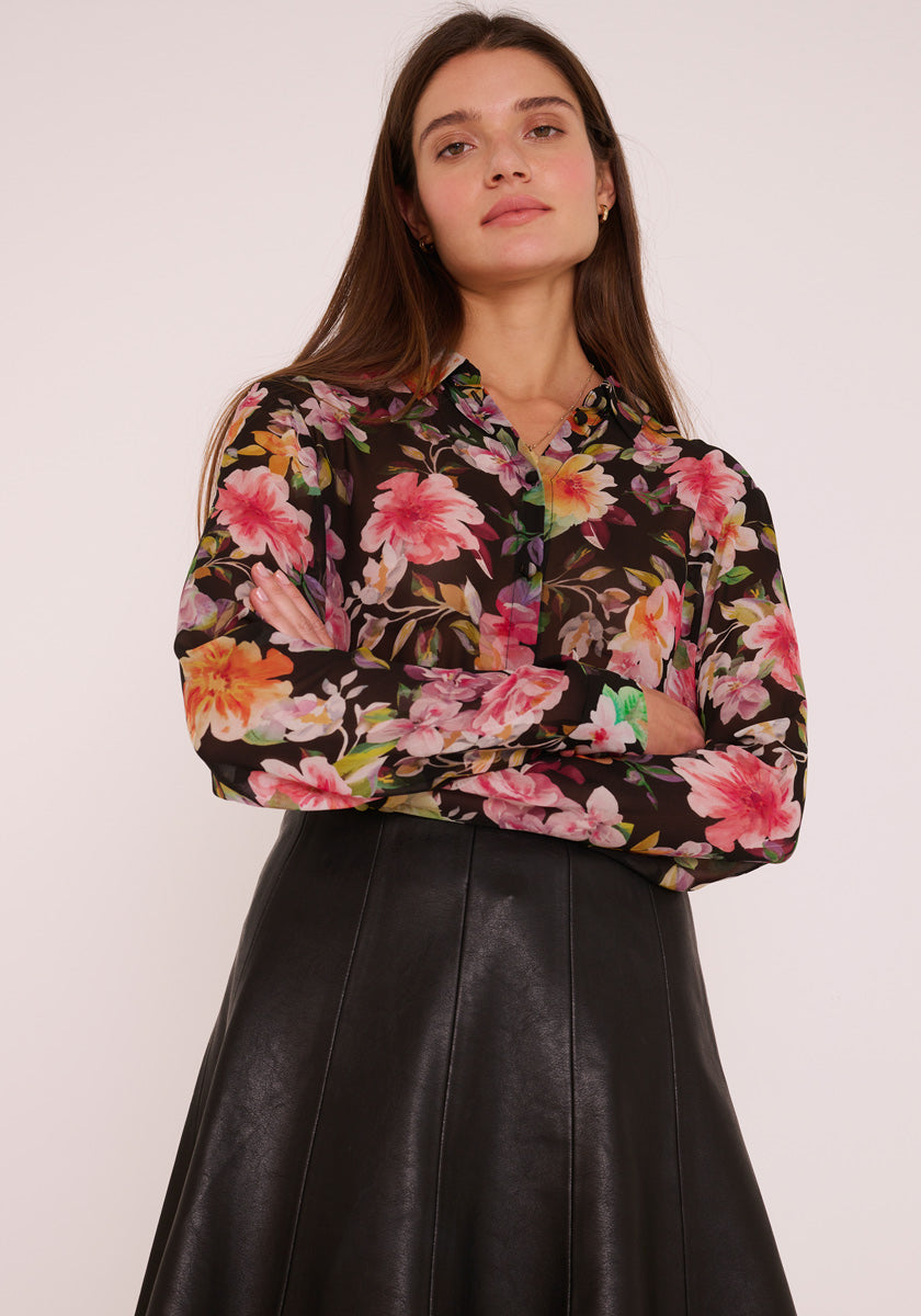 Eine Frau mit langen braunen Haaren steht selbstbewusst in einer schwarzen Princess Goes Hollywood-Hemdbluse mit lebhaftem rosa, orangefarbenem und grünem Blumenaufdruck, gepaart mit einem hochtaillierten schwarzen Kunstlederrock vor einem schlichten hellen Hintergrund.