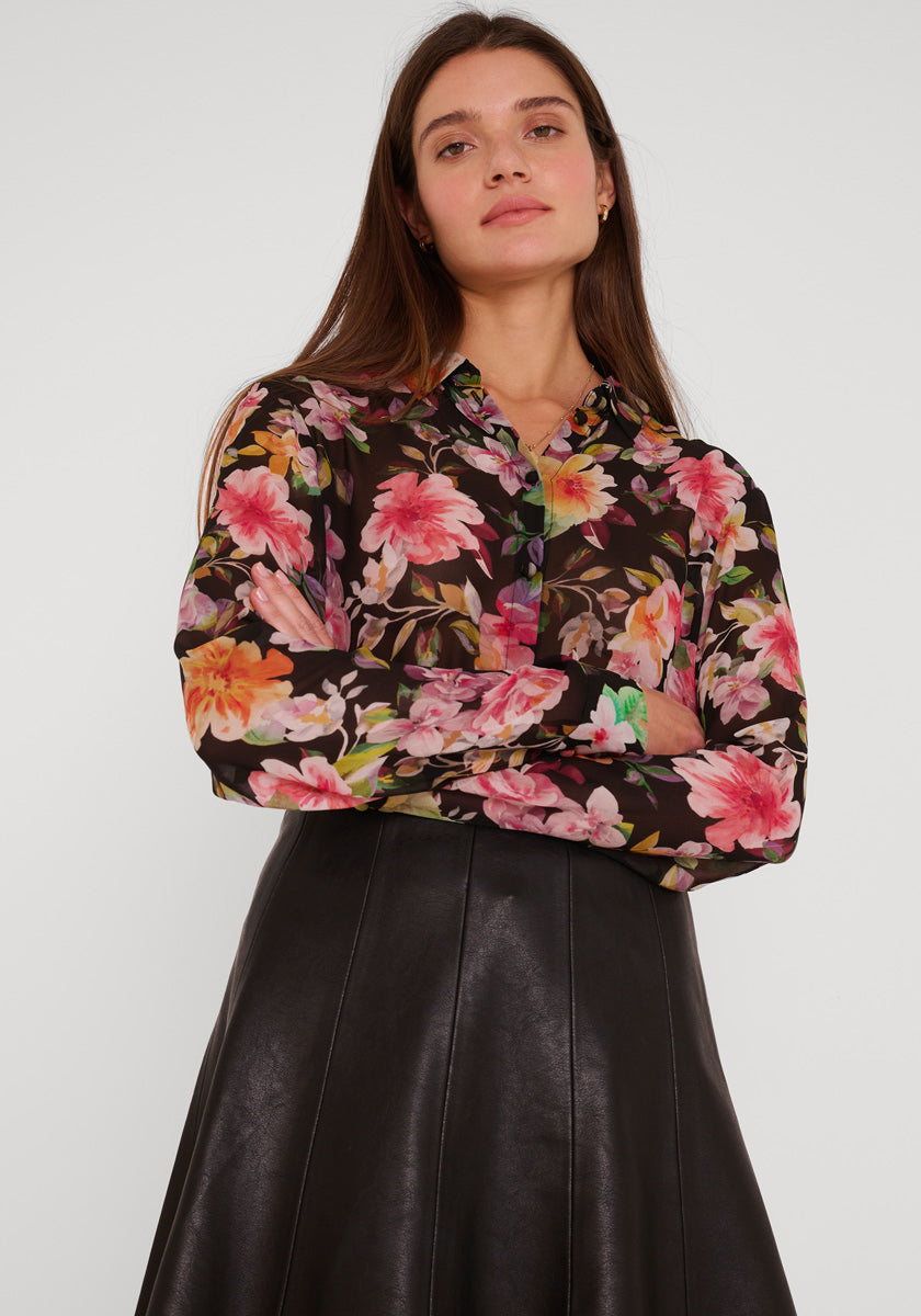 Eine Frau mit langen braunen Haaren steht selbstbewusst mit verschränkten Armen in einem schwarzen Kunstlederrock und einer Princess Goes Hollywood-Bluse mit Blumendruck und langen Ärmeln vor einem schlichten hellgrauen Hintergrund.