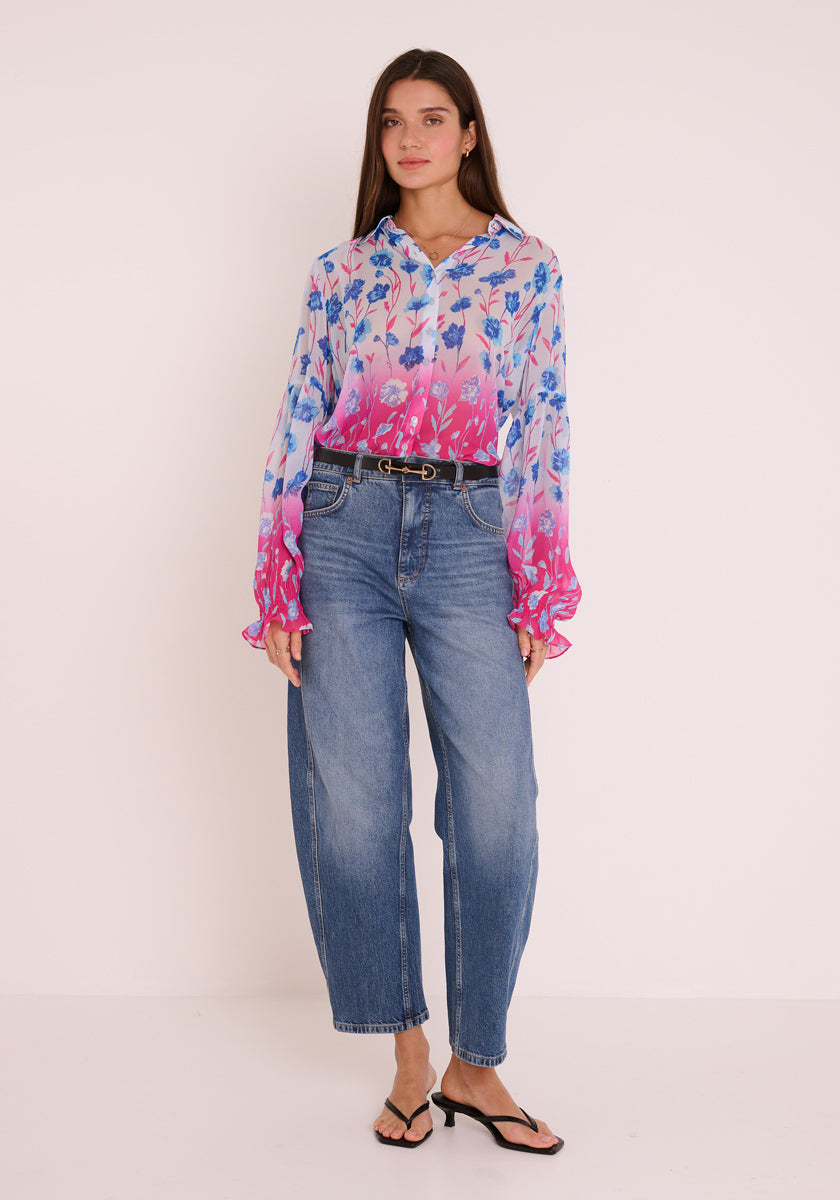 Eine Frau mit langen braunen Haaren trägt eine Princess Goes Hollywood-Hemdbluse mit Blumendruck in Blau- und Rosatönen, die in eine hoch taillierte Bluejeans mit schwarzem Gürtel gesteckt wird. Sie steht in schwarzen Sandalen vor einem hellen Hintergrund und wirkt entspannt und neutral.