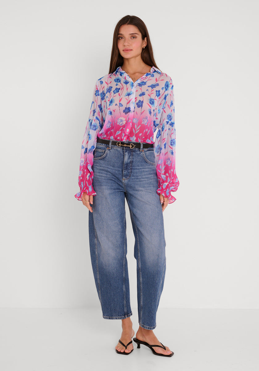 Eine Frau mit glattem braunem Haar steht vor einem weißen Hintergrund und trägt eine bunte Hemdbluse mit Blumendruck und langen Ärmeln von Princess Goes Hollywood, blaue High-Waisted-Jeans mit schwarzem Gürtel und schwarze Zehensandalen.