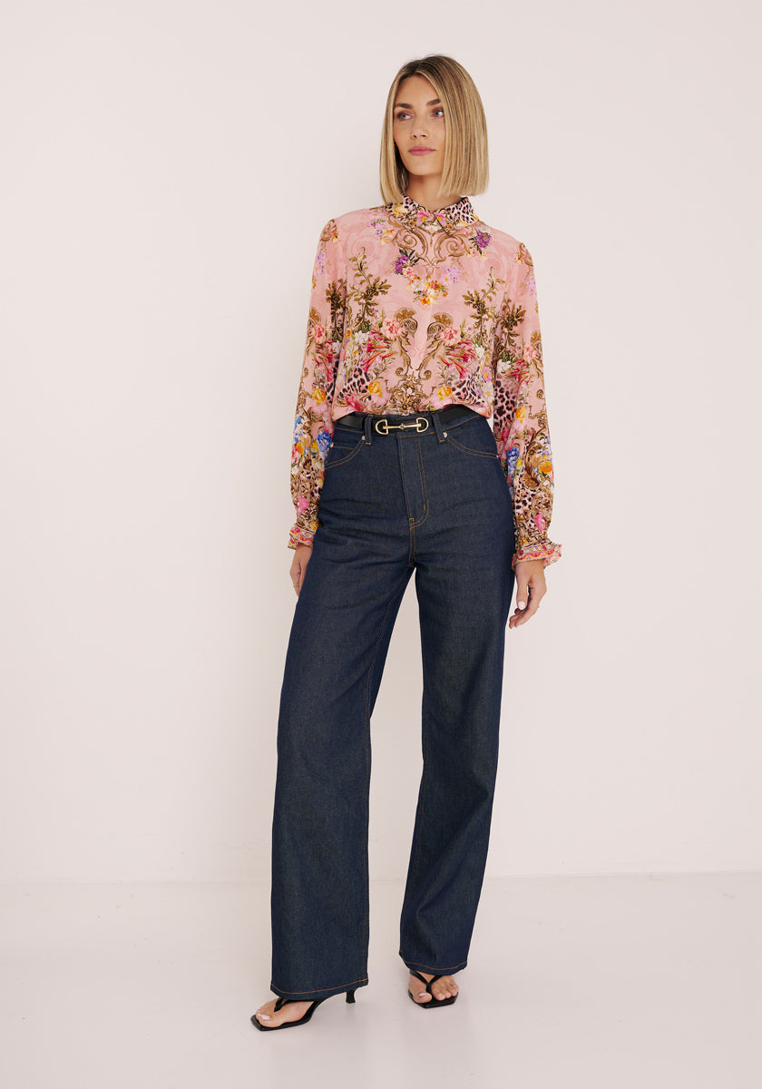 Eine Frau mit blondem Bob trägt eine rosa, langärmelige Seidenbluse mit Blumenaufdruck von Princess Goes Hollywood, gepaart mit einer dunkelblauen, weit geschnittenen Jeans mit hoher Taille, einem Gürtel und schwarzen hochhackigen Sandalen. Sie steht vor einem weißen Hintergrund.