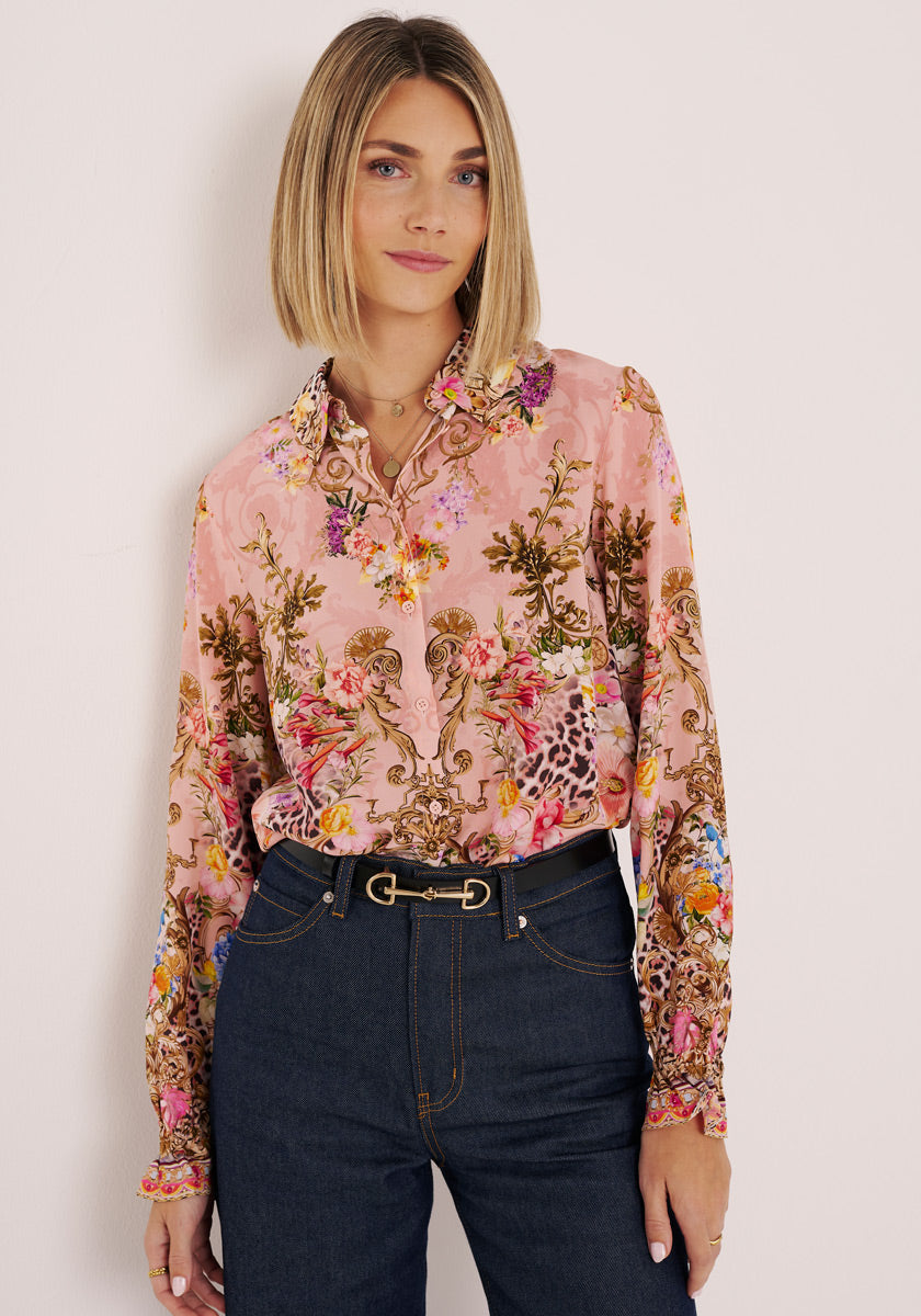 Eine Frau mit glattem, schulterlangem blondem Haar steht vor einer hellen Wand und trägt eine rosafarbene Seidenbluse mit Blumenmuster von Princess Goes Hollywood, die in eine dunkle Jeans mit hoher Taille und einem schmalen schwarzen Gürtel gesteckt wird. Sie blickt neutral in die Kamera.