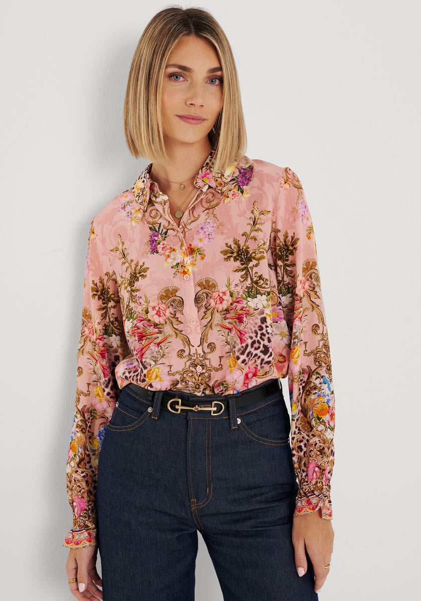 Eine Frau mit glattem, schulterlangem blondem Haar trägt eine rosafarbene Hemdbluse mit Blumendruck von Princess Goes Hollywood, die in eine dunkelblaue Jeans mit hoher Taille und einem schwarzen Gürtel gesteckt wird. Sie steht vor einer schlichten weißen Wand und blickt neutral in die Kamera.