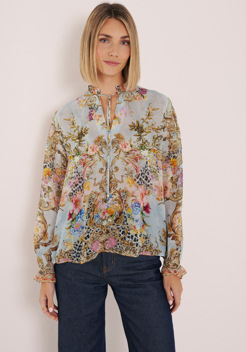 Eine Frau mit glattem, schulterlangem blondem Haar modelliert eine hellblaue Princess Goes Hollywood Seidenbluse mit einem komplizierten Blumenaufdruck in Rosa-, Gold- und Grüntönen, gepaart mit dunkelblauen Jeans vor einem schlichten hellen Hintergrund.