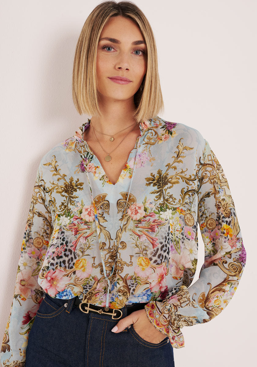 Eine Frau mit glattem blondem Haar und Seitenscheitel trägt eine helle, langärmelige Seidenbluse von Princess Goes Hollywood mit Blumen- und Goldmuster, goldene Halsketten, dunkle High-Waisted-Jeans und einen Gürtel, vor einem hellen Hintergrund.