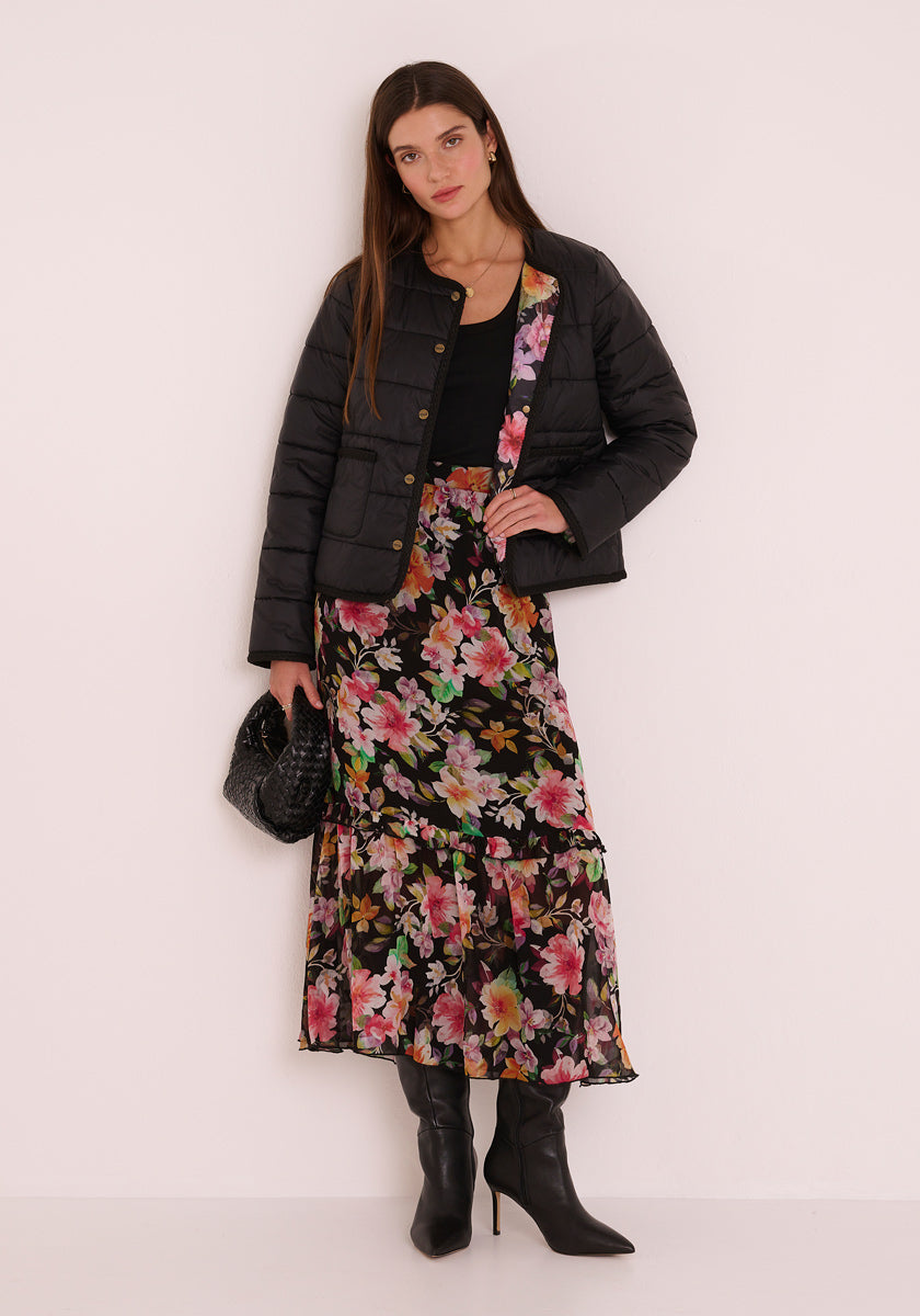 Eine Frau mit langen glatten braunen Haaren trägt eine schwarze Pufferjacke von Princess Goes Hollywood über einem schwarzen Oberteil und einem geblümten Midirock mit rosa und grünen Akzenten, dazu schwarze spitze Stiefel und eine schwarze Clutch mit Textur vor einer hellen Wand.