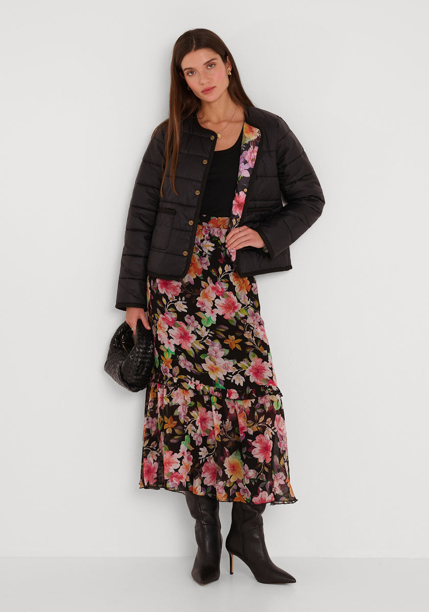 Eine Frau mit langen braunen Haaren und neutralem Gesichtsausdruck steht vor einer weißen Wand. Sie trägt eine schwarze Pufferjacke von Princess Goes Hollywood über einem schwarzen Top, einen langen Midirock mit Blumenmuster, schwarze spitze Stiefel und eine schwarze Handtasche mit Struktur.