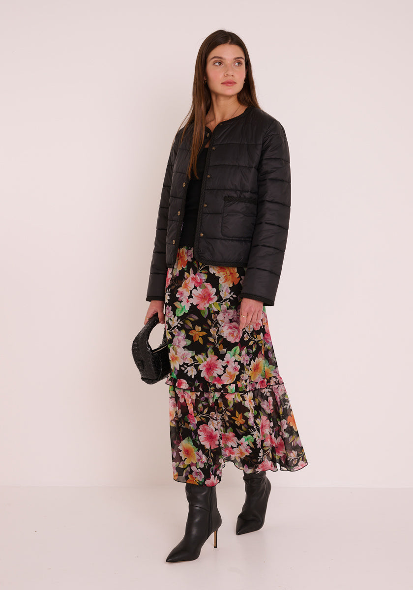Eine Frau steht vor einem schlichten Hintergrund und trägt eine schwarze Steppjacke, ein schwarzes Oberteil, einen Flower-Print-Midirock von Princess Goes Hollywood mit rosa, grünen und gelben Blumen, spitze schwarze Stiefel und eine kleine schwarze Tasche.