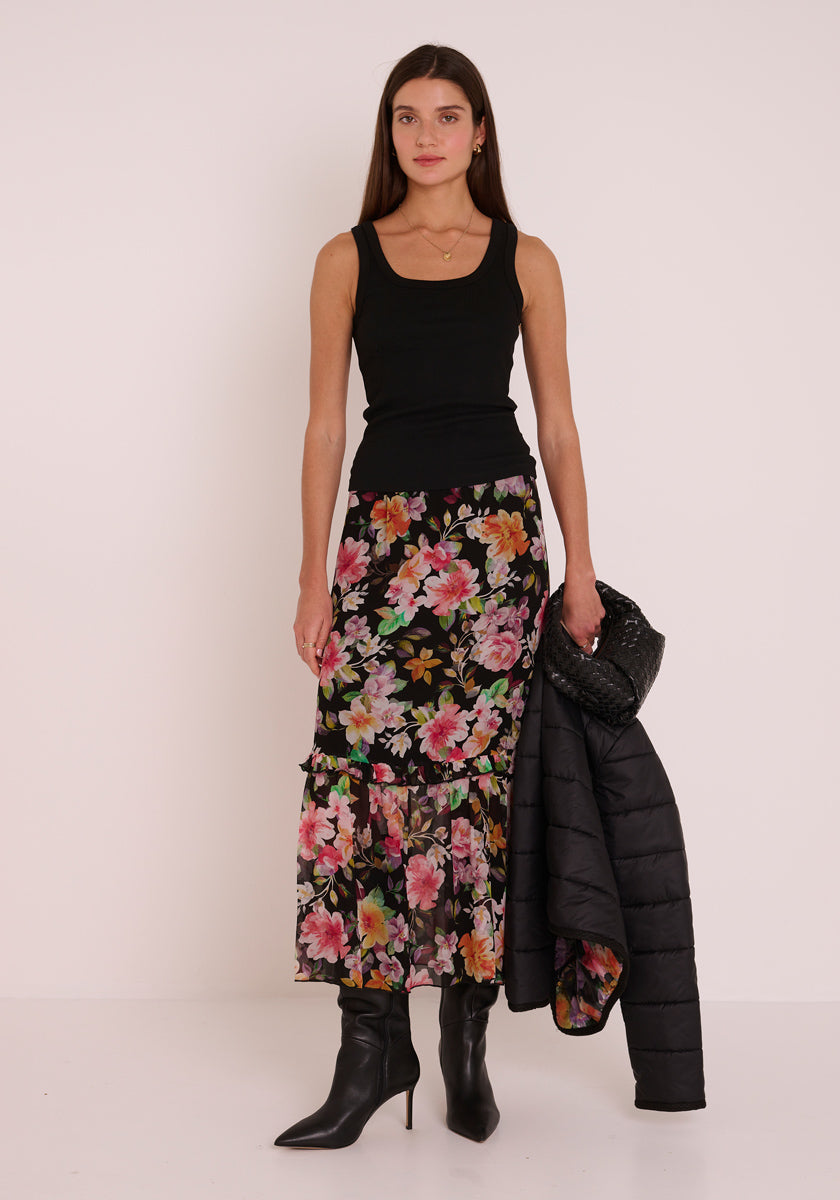 Eine Frau posiert in einem ärmellosen schwarzen Oberteil und einem geblümten Midirock von Princess Goes Hollywood vor einer schlichten hellen Wand. Sie trägt eine schwarze Steppjacke und eine Handtasche und vervollständigt den Look mit schwarzen Stiefeln mit spitzen Absätzen.