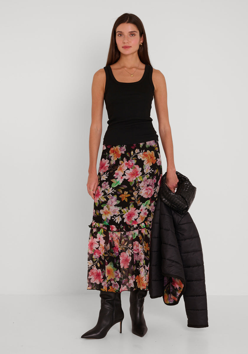 Eine Frau mit langen glatten braunen Haaren trägt ein schwarzes ärmelloses Rundhalsausschnitt-Top, einen langen schwarzen geblümten Rock und schwarze hochhackige Stiefel von Princess Goes Hollywood. Sie hält eine gesteppte Kurzjacke in ihrer linken Hand vor einem weißen Hintergrund.