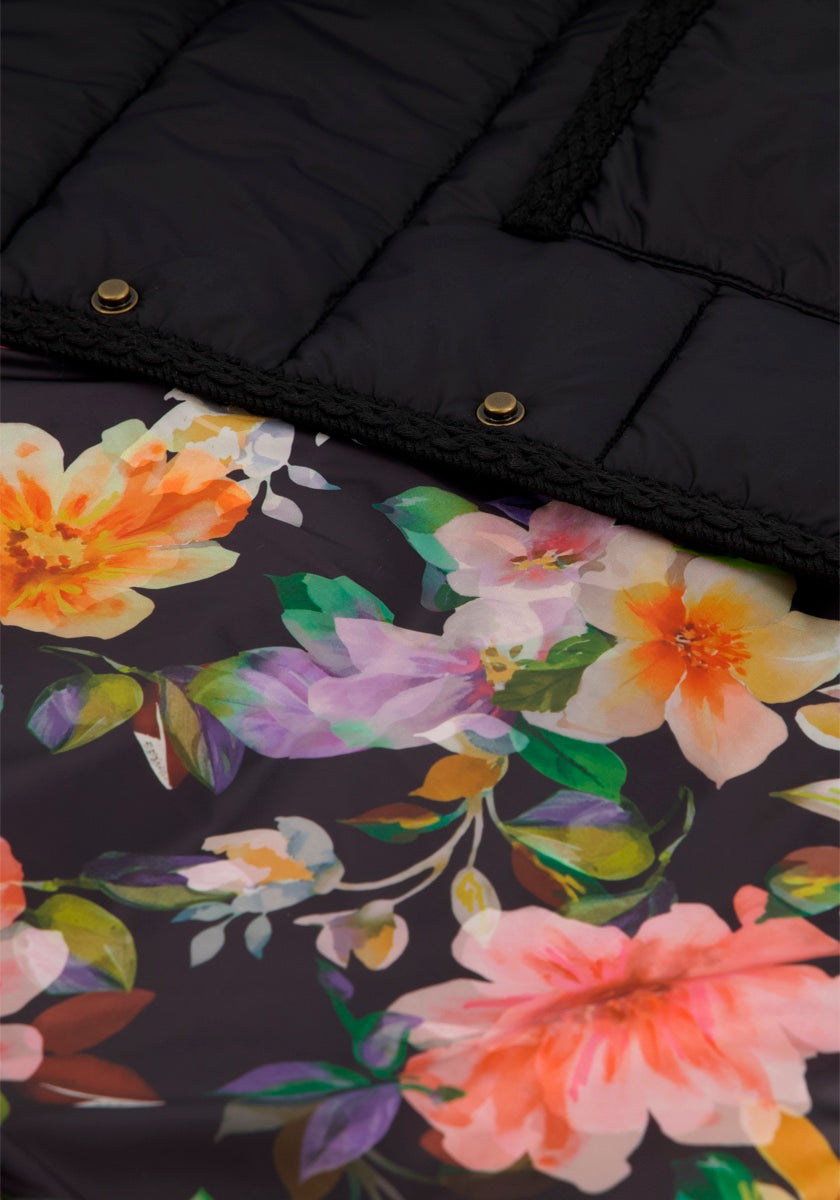 Nahaufnahme einer Princess Goes Hollywood Gesteppte Kurzjacke mit leuchtend rosa, orange, gelb und grünem floralem Innenfutter. Dunkler gesteppter Stoff mit zwei metallischen Druckknöpfen und schwarzen Nähten sitzt über dem geblümten Innenfutter.