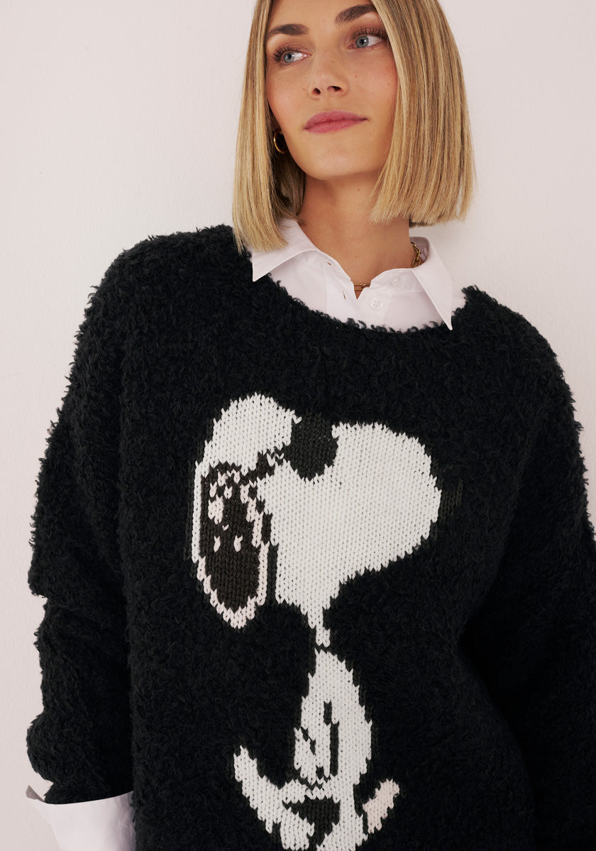 Eine Frau mit glattem, schulterlangem blondem Haar trägt einen Princess Goes Hollywood Kuschelpullover aus Wollmix mit handgestricktem Snoopy-Motiv über einer weißen Bluse. Sie lächelt sanft und blickt leicht zur Seite vor schlichtem Hintergrund.
