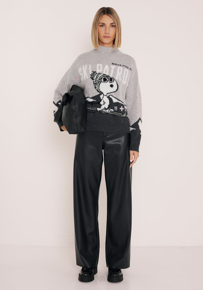 Eine Frau mit stumpfem Haarschnitt steht vor einer schlichten Wand. Sie trägt einen grauen Wollpullover mit Snoopy-Motiv, eine schwarze Lederhose mit weitem Bein, klobige schwarze Schuhe und eine große schwarze Clutch. Sie blickt neutral nach vorne.