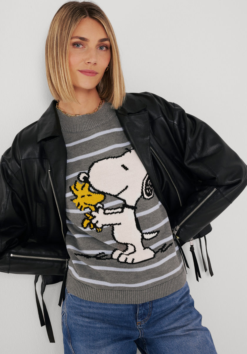 Eine Frau mit glattem blondem Haar trägt eine schwarze Lederjacke von Princess Goes Hollywood über einem grauen Snoopy-hugging-Woodstock Baumwollmix-Pullover, gepaart mit blauen Jeans und einer goldenen Halskette, vor einem schlichten weißen Hintergrund.