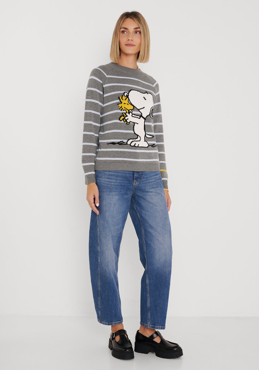 Eine Frau mit schulterlangem blondem Haar steht vor einer weißen Wand. Sie trägt einen Princess Goes Hollywood Gestreifter Pullover mit Snoopy &amp; Woodstock zum Umarmen, weichen Baumwollmix, weite Bluejeans und schwarze Schuhe im Mary-Jane-Stil. Sie blickt in die Kamera.
