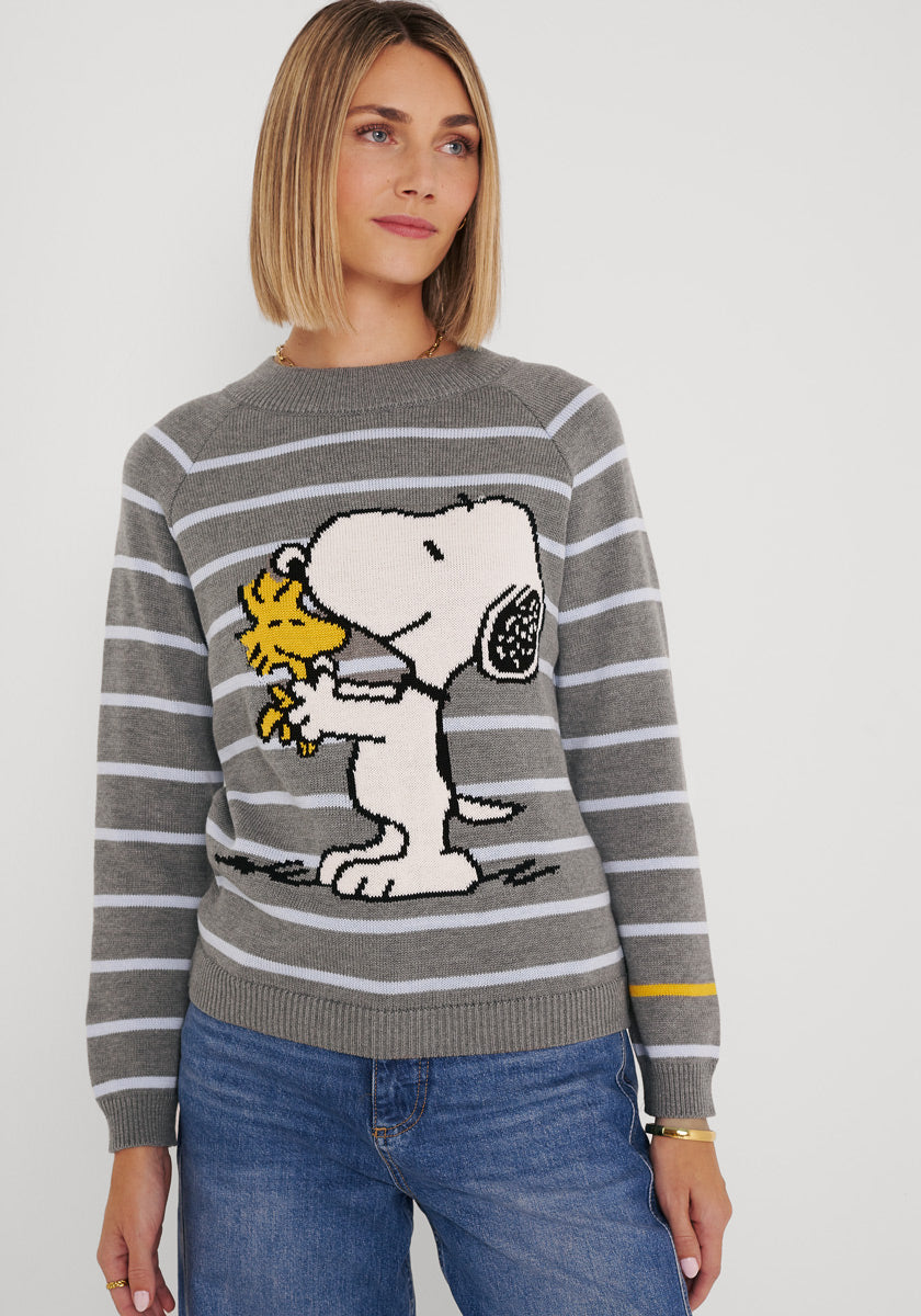 Eine Frau mit glattem blondem Haar trägt einen gestreiften Baumwollmischpullover von Princess Goes Hollywood, auf dessen Vorderseite sich Snoopy und Woodstock umarmen. Sie steht in blauen Jeans vor einem weißen Hintergrund, ihr Gesichtsausdruck ist neutral.