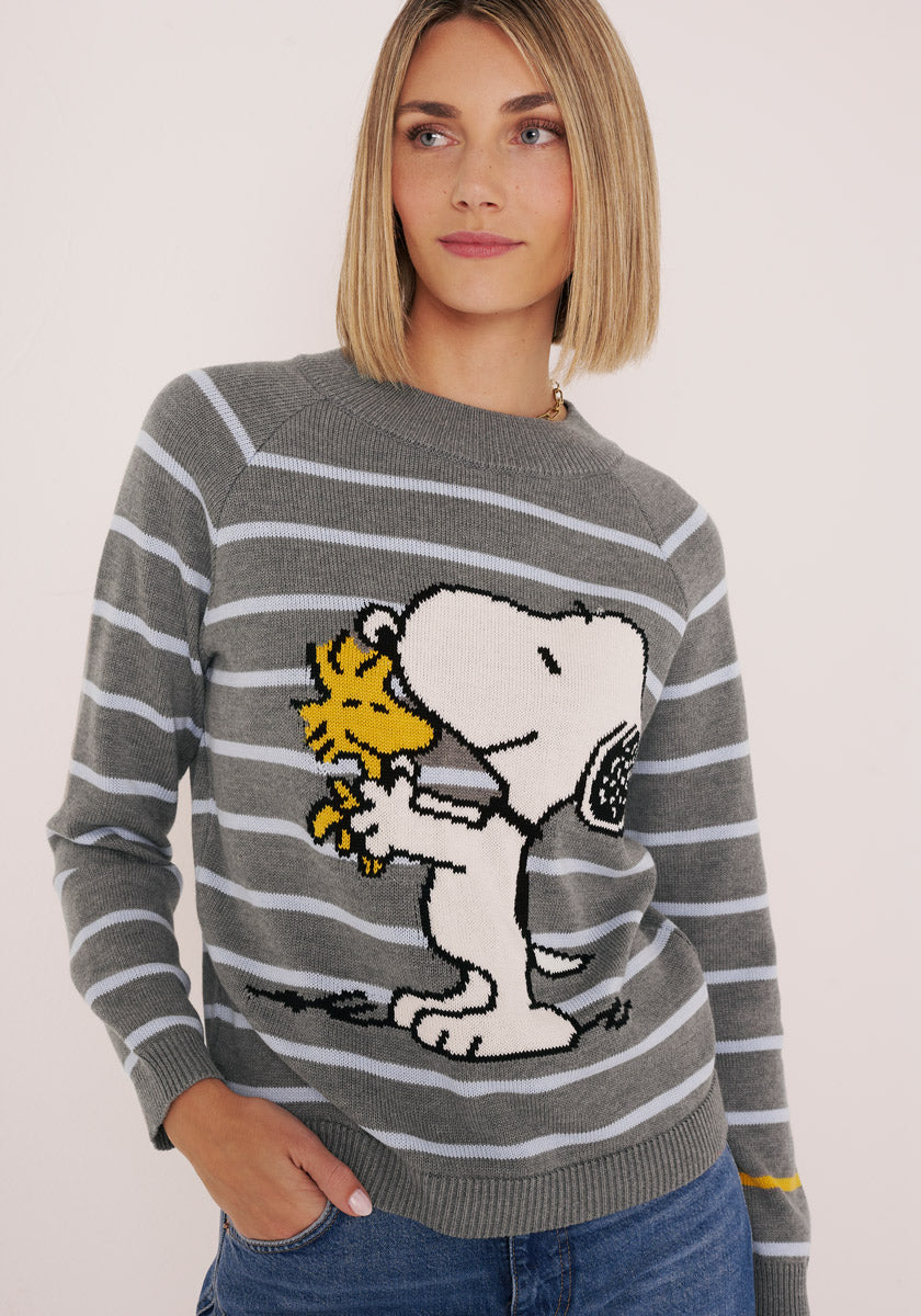 Eine Frau mit glattem blondem Haar trägt einen gestreiften Baumwollpullover von Princess Goes Hollywood, auf dem eine große Illustration von Snoopy zu sehen ist, der Woodstock umarmt. Sie steht mit einer Hand in ihrer Jeanstasche und lächelt sanft zur Seite.