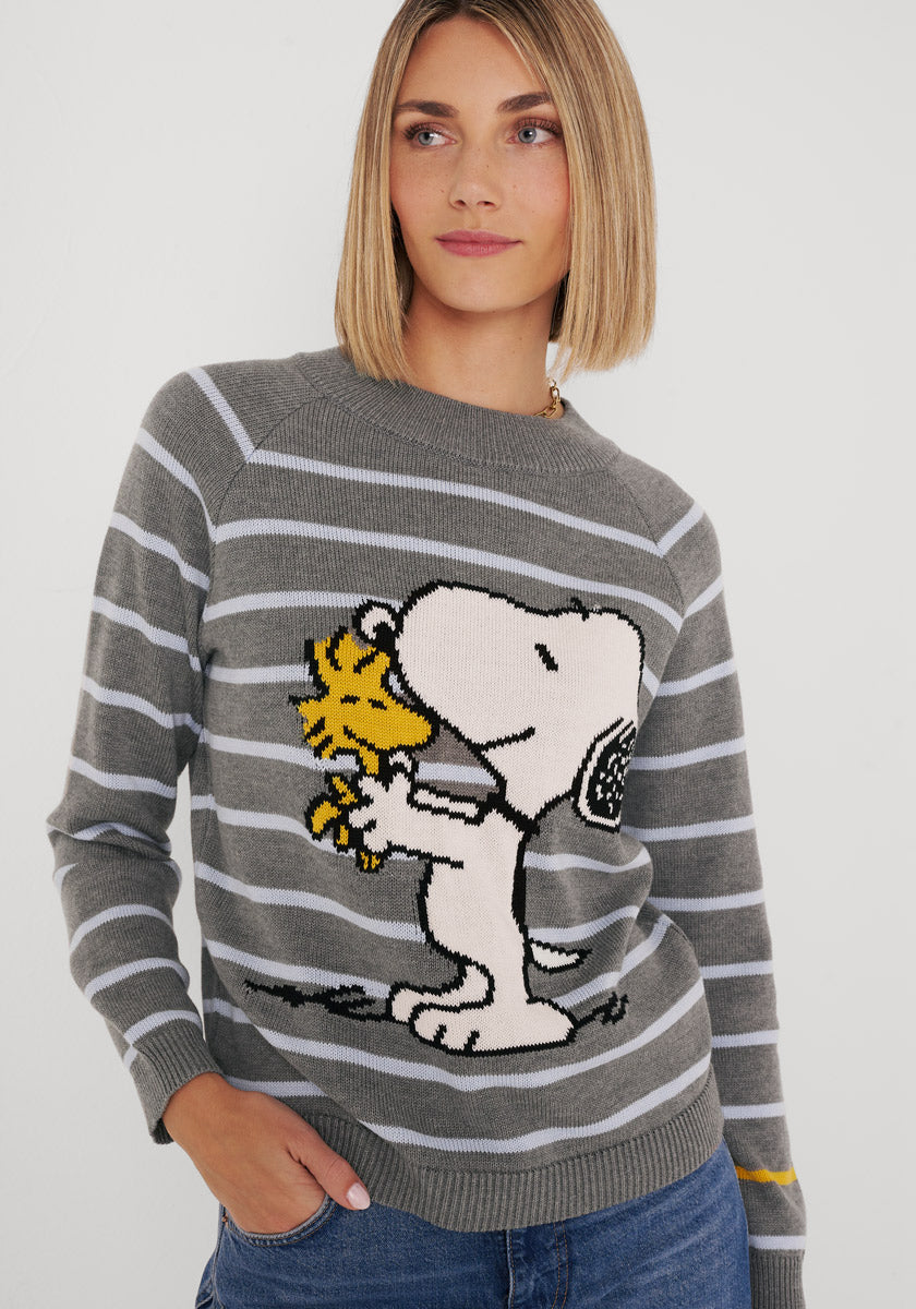 Eine Frau mit glattem blondem Haar trägt einen Princess Goes Hollywood gestreifter Baumwollmix-Pullover mit großem Snoopy-Woodstock-Print. Sie lächelt sanft, steht vor einem weißen Hintergrund und hat eine Hand in ihrer Jeanstasche.