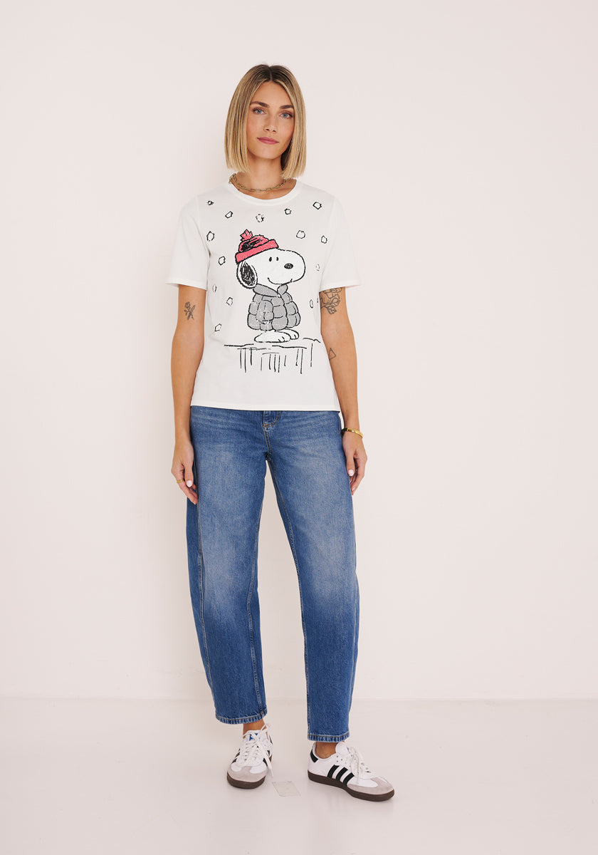 Eine Frau mit einem stumpfen Bob steht vor einer weißen Wand. Sie trägt ein Princess Goes Hollywood Snoopy T-Shirt aus Baumwolle, blaue Jeans und weiße Turnschuhe mit schwarzen Streifen. Ihre Arme liegen entspannt an ihren Seiten, während sie in die Kamera schaut.