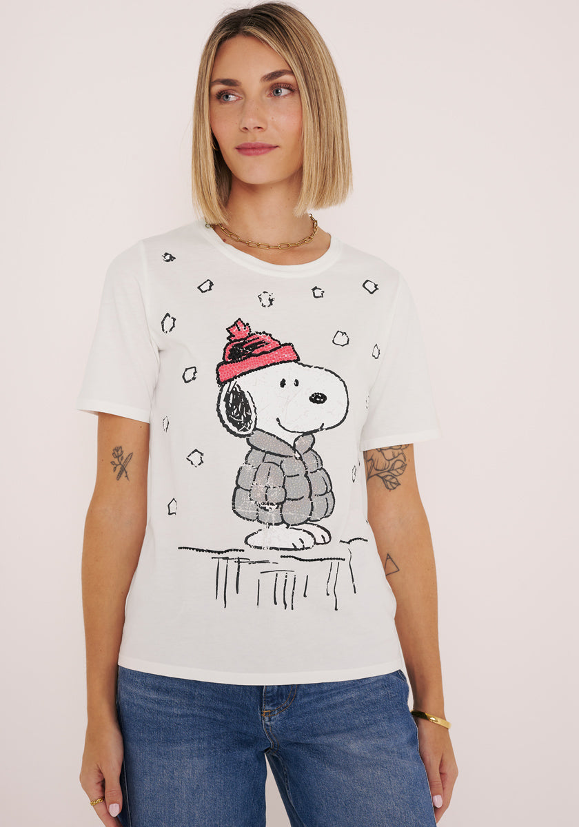 Eine Frau mit Tattoos trägt ein weißes Princess Goes Hollywood Snoopy T-Shirt aus Baumwolle, auf dem Snoopy mit roter Mütze und Pufferjacke im Schnee abgebildet ist, gepaart mit blauen Jeans und Schmuck, vor einem blassrosa Hintergrund.