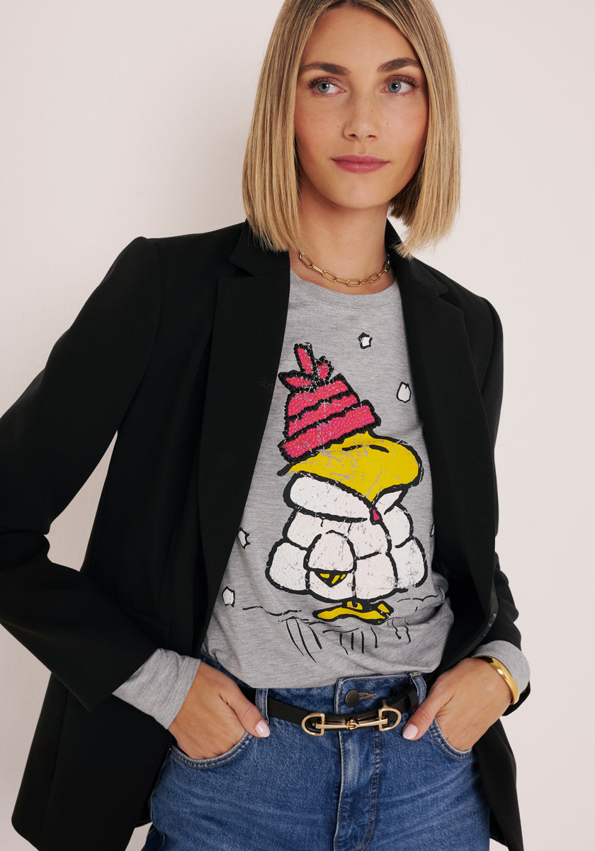 Eine Frau mit glattem, schulterlangem blondem Haar trägt einen schwarzen Blazer über einem grauen, langärmeligen Princess Goes Hollywood-Shirt mit Woodstock-Aufdruck. Sie steht vor einem schlichten Hintergrund und blickt leicht zur Seite, ruhig und gelassen.