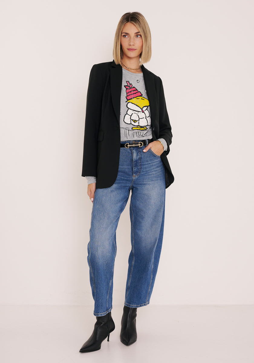 Eine Frau mit blondem Bob steht vor einer weißen Wand. Sie trägt einen schwarzen Blazer von Princess Goes Hollywood über einem grauen langärmeligen Hemd mit einem Cartoon-Huhn in einem Partyhut, blaue Jeans mit hoher Taille, einen schwarzen Gürtel mit goldener Schnalle und schwarze Ankle Boots. Linke Hand in der Tasche.