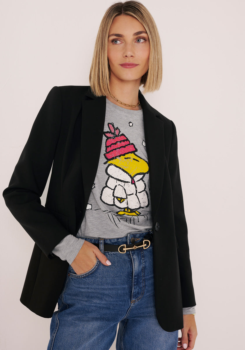 Eine Frau mit glattem blondem Bob trägt einen schwarzen Blazer über einem grauen Princess Goes Hollywood Langarmshirt mit Woodstock-Print, auf dem eine Cartoon-Ente mit rosa Mütze und Pufferjacke abgebildet ist, gepaart mit blauen Jeans, vor einem schlichten hellen Hintergrund.