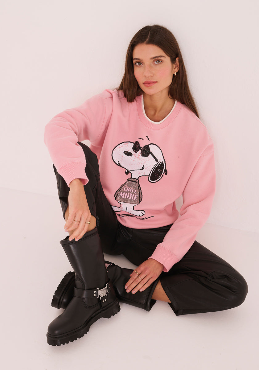 Eine Frau mit langen braunen Haaren sitzt vor einer weißen Wand, trägt ein pinkfarbenes Princess Goes Hollywood Sweatshirt Damen mit Snoopy-Print und "Chill More", eine schwarze Lederhose und schwarze Stiefel und blickt in die Kamera, einen Arm auf ihr Knie gestützt.