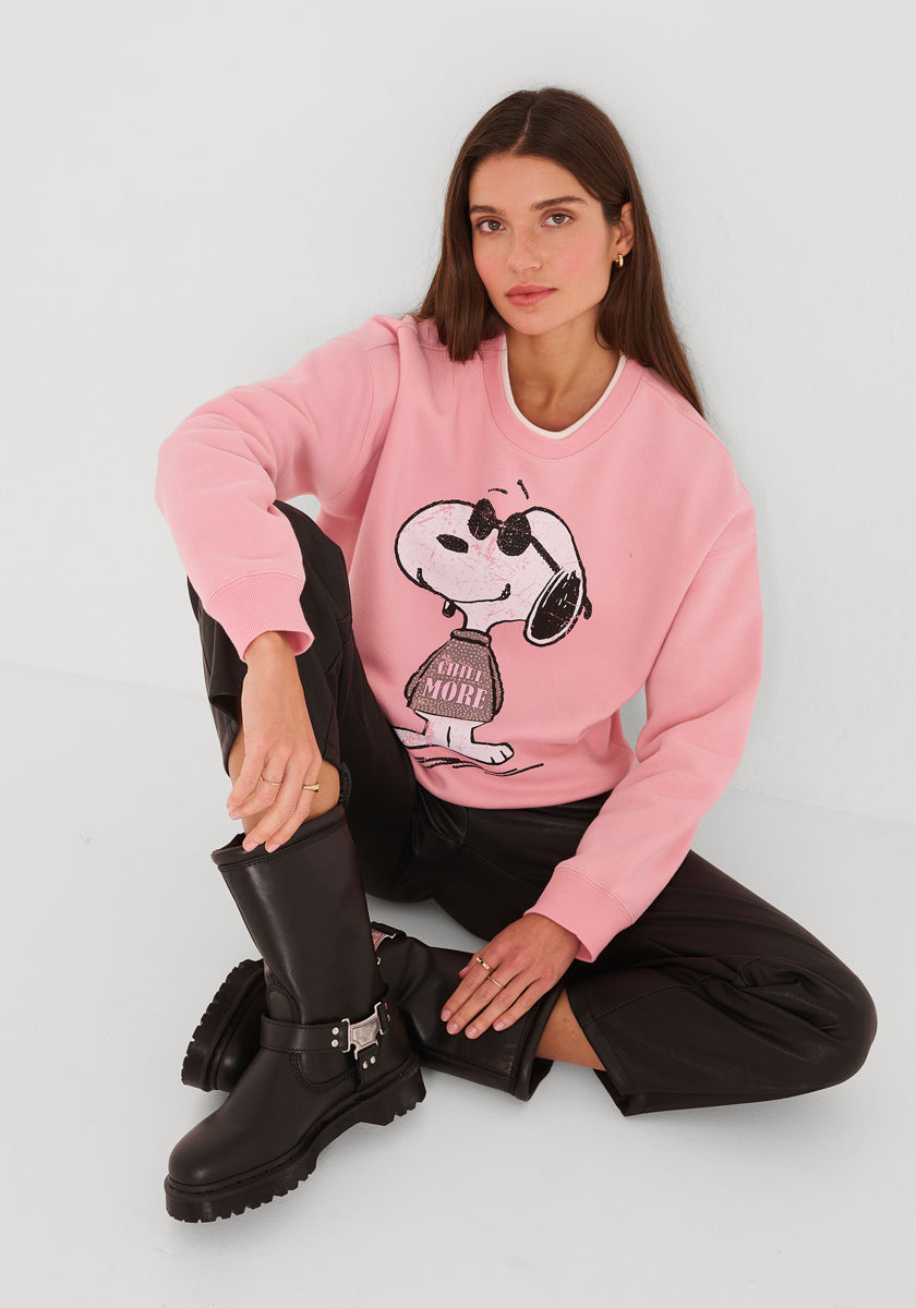 Eine Frau mit langen braunen Haaren sitzt vor einer weißen Wand und trägt ein Princess Goes Hollywood Sweatshirt Damen mit einem coolen Snoopy-Aufdruck, eine schwarze Kunstlederhose und klobige schwarze Stiefel. Sie schaut entspannt in die Kamera, einen Arm auf ihr Knie gestützt.