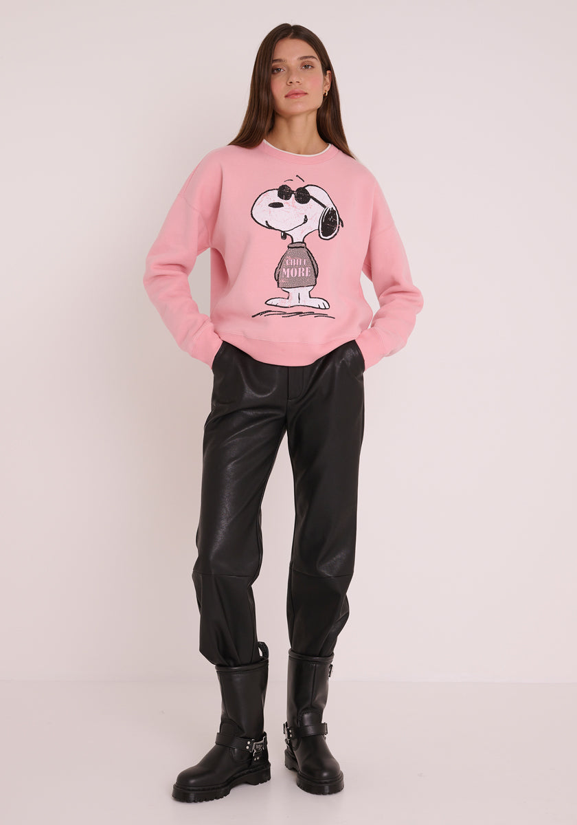 Eine Frau steht vor einer schlichten weißen Wand in einem hellrosa Princess Goes Hollywood Sweatshirt Damen mit coolem Snoopy-Print, schwarzer Kunstlederhose und schwarzen Schnallenstiefeln. Die Hände in den Taschen, schaut sie selbstbewusst in die Kamera.