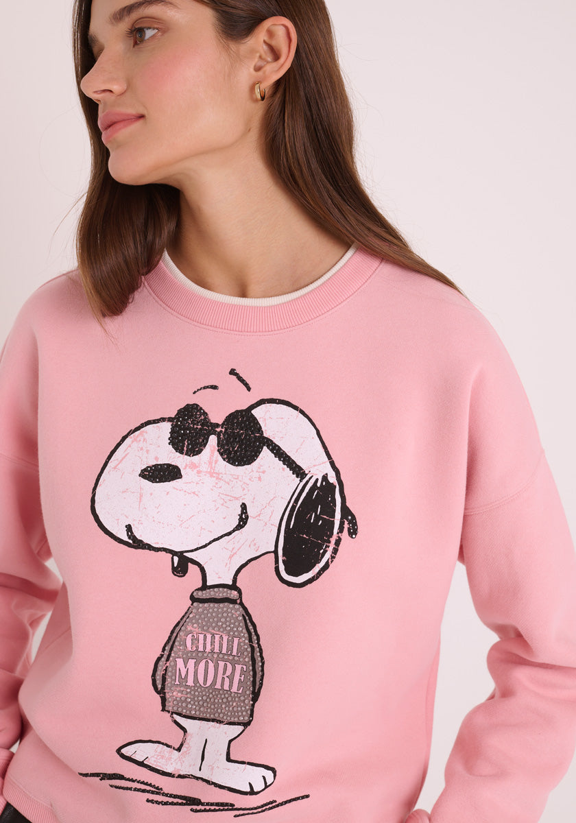 Eine Frau mit glattem braunem Haar trägt ein rosa Princess Goes Hollywood Sweatshirt Damen mit einem Snoopy mit Sonnenbrille und einem Pullover mit der Aufschrift &quot;CHILL MORE&quot;. Sie steht vor einem schlichten hellen Hintergrund, schaut leicht nach links und lächelt sanft.