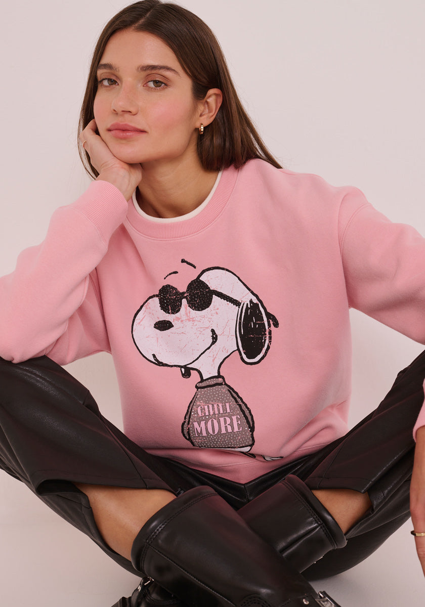 Eine Frau mit glattem braunem Haar sitzt im Schneidersitz und stützt ihr Gesicht auf ihre Hand. Sie trägt ein rosafarbenes Princess Goes Hollywood Sweatshirt Damen mit Snoopy-Print und &quot;CHILL MORE&quot;-Design, gepaart mit einer schwarzen Hose und kniehohen Stiefeln.