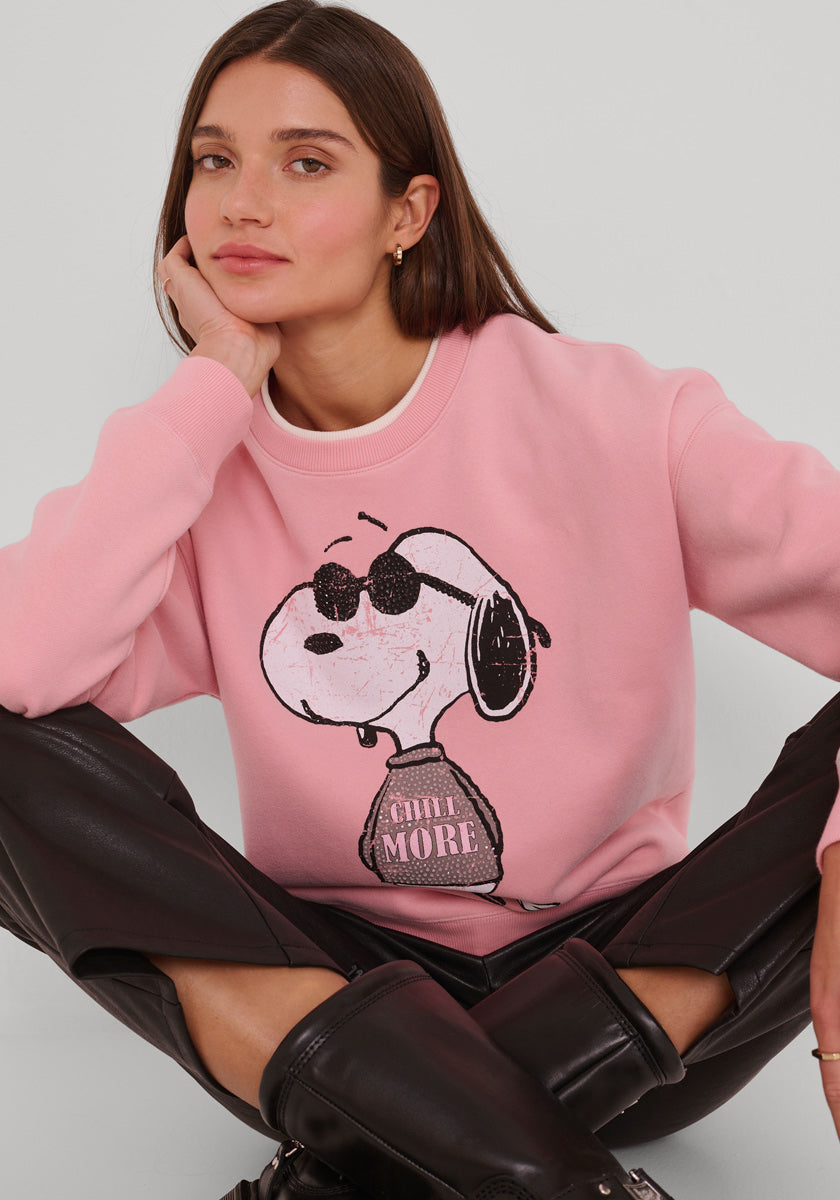 Eine Frau mit glattem braunem Haar trägt ein rosa Princess Goes Hollywood Snoopy-Print Sweatshirt Damen mit Snoopy mit Sonnenbrille und "CHILL MORE", gepaart mit einer schwarzen Lederhose und Stiefeln, vor einem schlichten grauen Hintergrund.
