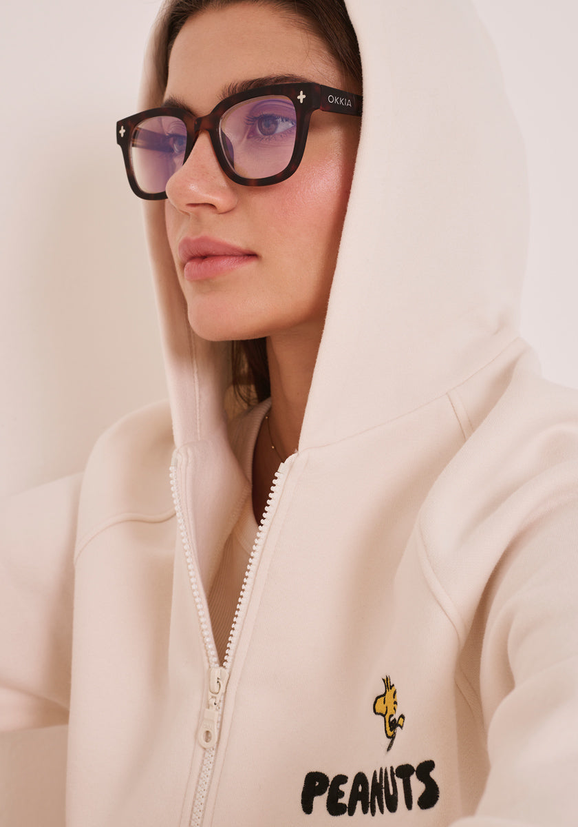 Eine Frau mit langen braunen Haaren und schwarzer Brille trägt einen cremefarbenen Princess Goes Hollywood Peanuts-Kapuzenpullover mit aufgesetzter Kapuze. Auf der Brust ist ein Woodstock und &quot;PEANUTS&quot; aufgestickt. Sie blickt leicht nach links vor einem einfarbigen Hintergrund.