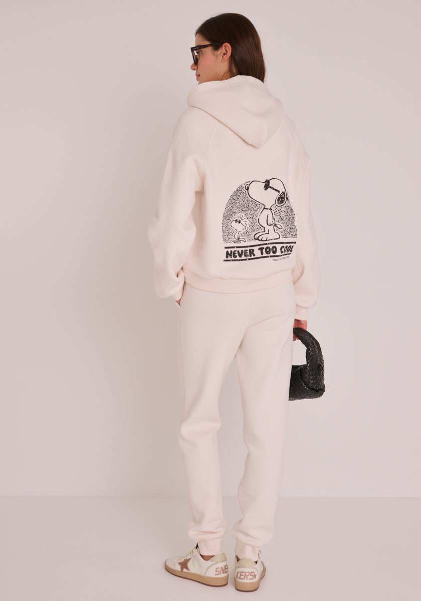 Eine Frau in einem cremefarbenen Princess Goes Hollywood-Sweat-Hoodie mit Peanuts-Stickerei und einem auffälligen Snoopy-Aufdruck auf dem Rücken, einer passenden Jogginghose, beigen Turnschuhen, einer Sonnenbrille und einer strukturierten schwarzen Handtasche vor einem weißen Hintergrund.