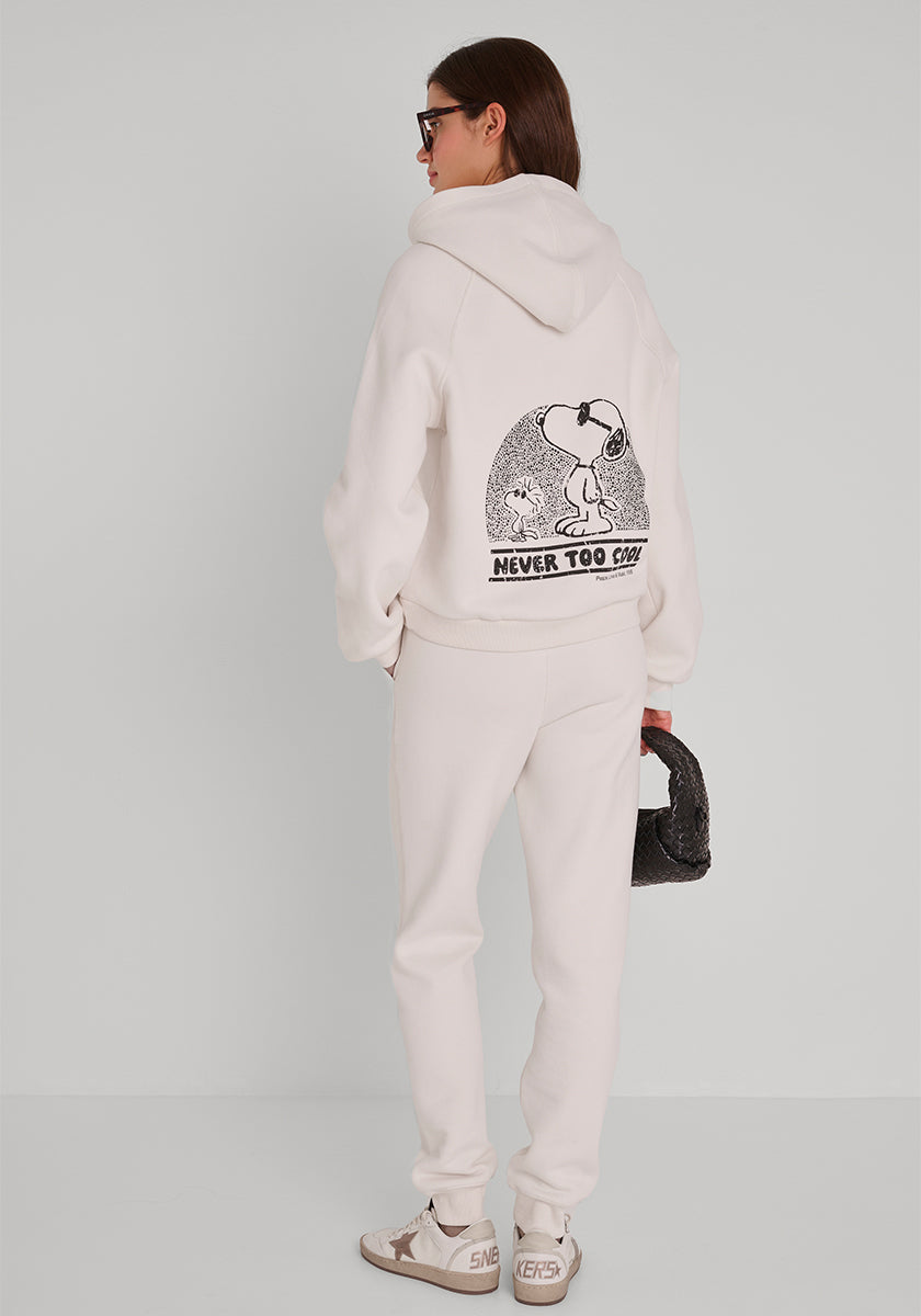 Eine Frau mit braunen Haaren trägt einen cremefarbenen Princess Goes Hollywood Baumwollmix-Kapuzenpullover mit einer großen Snoopy-Grafik und dem Text "Never Too Cool" auf dem Rücken, eine passende Sweathose, beigefarbene Turnschuhe, eine Sonnenbrille und eine schwarze Handtasche mit Struktur.