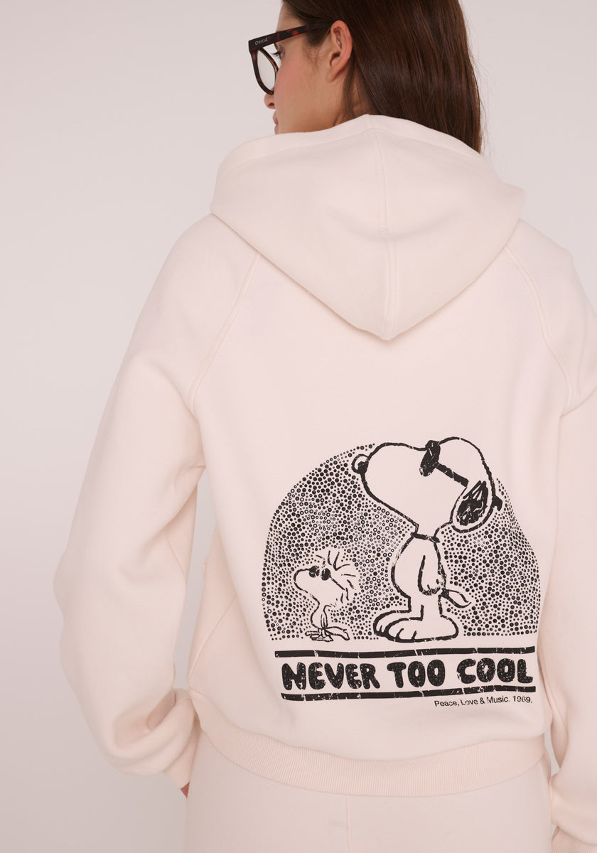 Eine Person mit langen braunen Haaren und Brille ist von hinten zu sehen. Sie trägt einen cremefarbenen Princess Goes Hollywood Sweat-Hoodie mit einem schwarz-weißen Snoopy-Aufdruck mit Woodstock und dem Text NEVER TOO COOL unter der Peanuts-Stickerei.