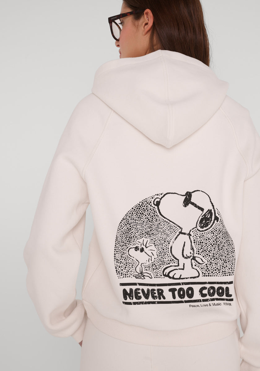 Eine Person mit langen braunen Haaren, die eine Brille trägt, steht in einem cremefarbenen Princess Goes Hollywood Peanuts-Kapuzenpullover mit einer schwarzen Linie als Snoopy- und Woodstock-Motiv auf dem Rücken, einem gepunkteten Halbkreis darüber und dem Text "NEVER TOO COOL" darunter.