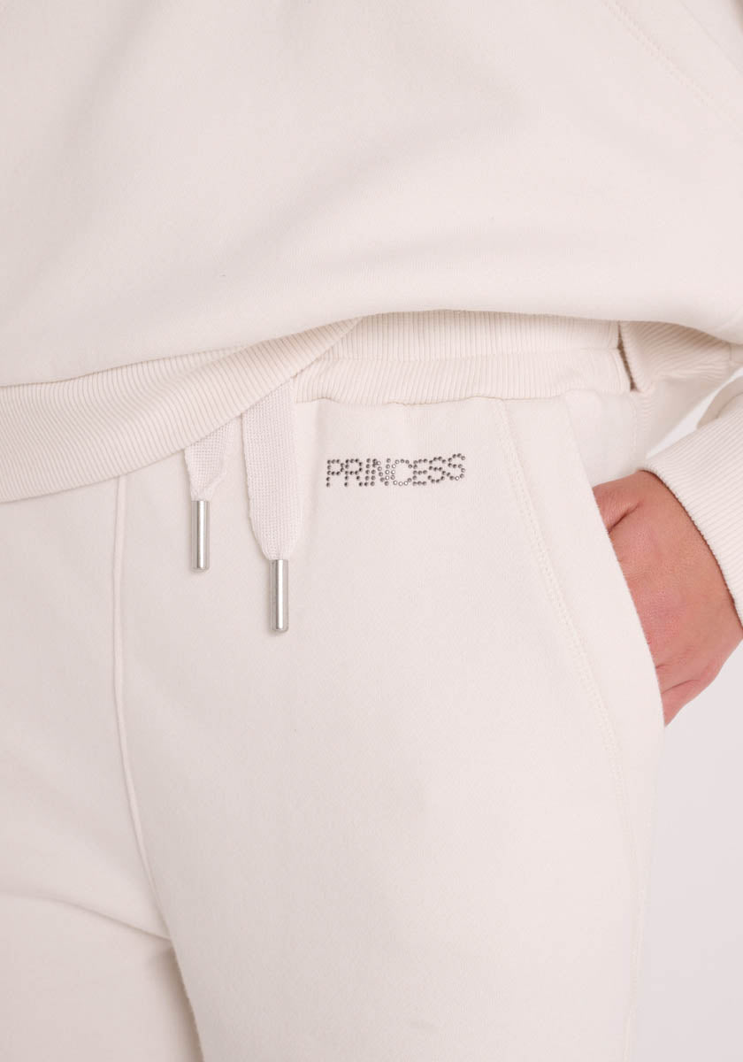 Sie tragen eine cremefarbene Sweathose und ein passendes Sweatshirt von Princess Goes Hollywood, ihre rechte Hand steckt in der Tasche. Der silberne PRINCESS-Schriftzug mit Strasssteinen akzentuiert den Bund, der durch Kordelzüge mit Metallspitzen in dieser kuscheligen Baumwollmischung ergänzt wird.