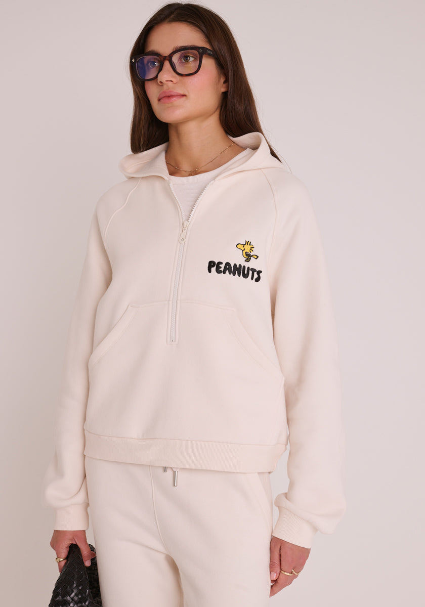 Eine Frau mit langen braunen Haaren und Brille trägt einen cremefarbenen Peanuts-Kapuzenpullover mit halbem Reißverschluss, der mit Snoopy&#39;s Woodstock und PEANUTS-Text auf der Brust bestickt ist, dazu eine schwarze Handtasche mit Struktur.