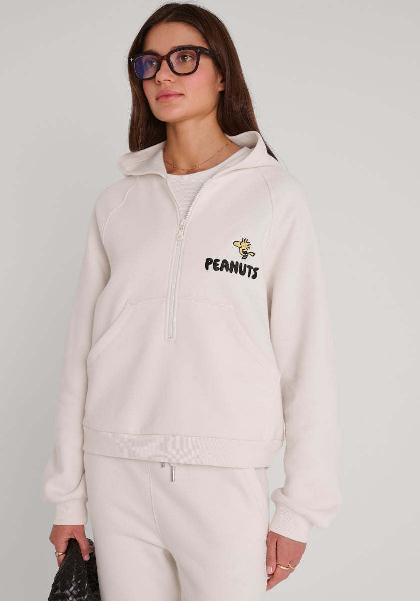 Eine Frau mit langen braunen Haaren und Brille trägt einen cremefarbenen Princess Goes Hollywood Peanuts-Kapuzenpulli mit einer kleinen Woodstock-Grafik und "PEANUTS" auf der Brust, dazu eine passende Hose und eine strukturierte schwarze Tasche vor einem schlichten Hintergrund.