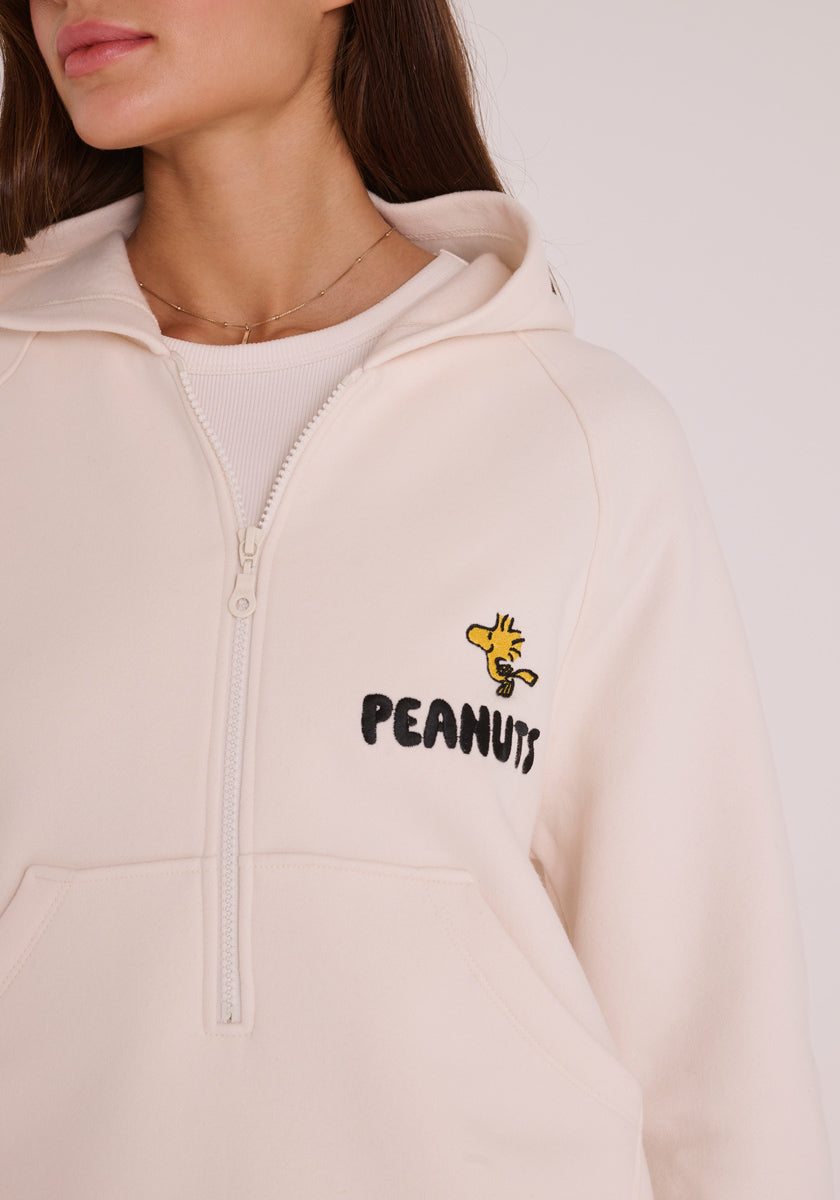 Eine Frau mit hellbraunem Haar trägt einen cremefarbenen Princess Goes Hollywood Peanuts Hoodie mit einem gestickten Woodstock über &quot;PEANUTS&quot; in schwarzer Schrift auf der linken Brust. Sie blickt leicht nach links und ist teilweise sichtbar. Perfekt für Fans der Comics.