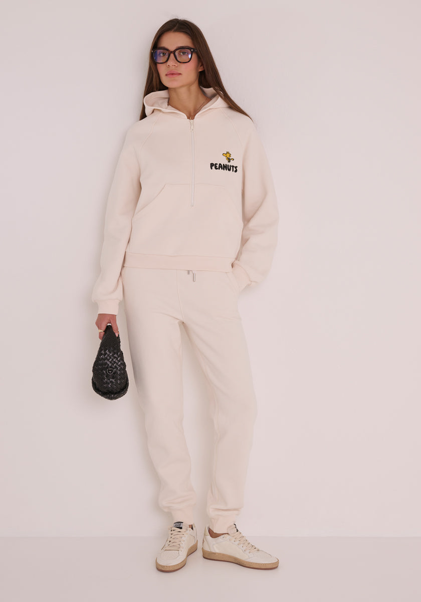 Eine Frau in einem cremefarbenen Princess Goes Hollywood-Sweat-Hoodie mit Peanuts-Stickerei und passender Jogginghose kombiniert den Look mit beigen Turnschuhen, einer übergroßen Sonnenbrille und einer kleinen schwarzen, strukturierten Handtasche und steht vor einer schlichten, hellen Wand.