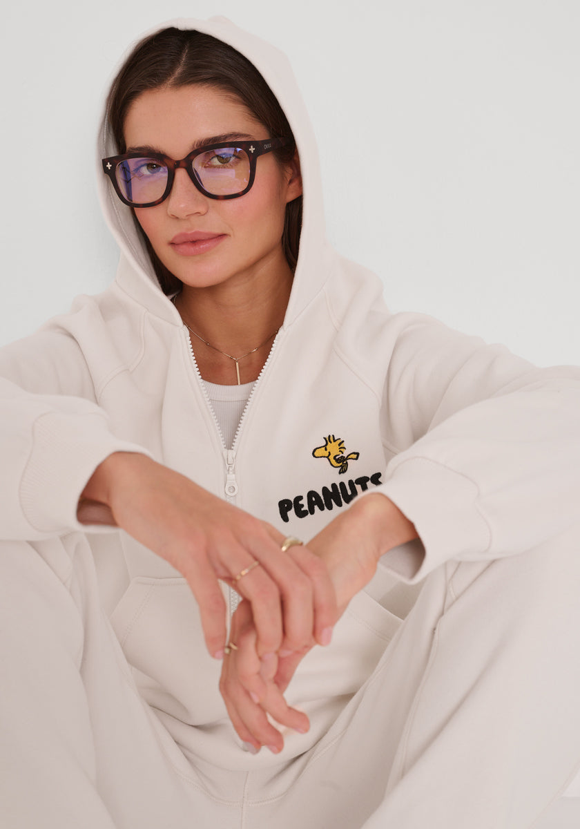 Eine Frau in einem cremefarbenen Princess Goes Hollywood-Peanuts-Kapuzenpullover mit dem Logo und dem Woodstock-Cartoon sitzt mit aufgesetzter Kapuze, Brille mit dunklem Gestell, hellbraunem Haar, die Hände zwischen den Knien, ruhig in die Kamera blickend vor einem schlichten Hintergrund.