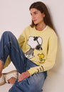 Eine Frau mit langen braunen Haaren sitzt in blauen Jeans und weißen Turnschuhen vor einer weißen Wand und trägt ein pastellgelbes Princess Goes Hollywood Snoopy Baumwoll-Sweatshirt mit Woodstock und "NEVER TOO COOL" auf dem Ärmel.