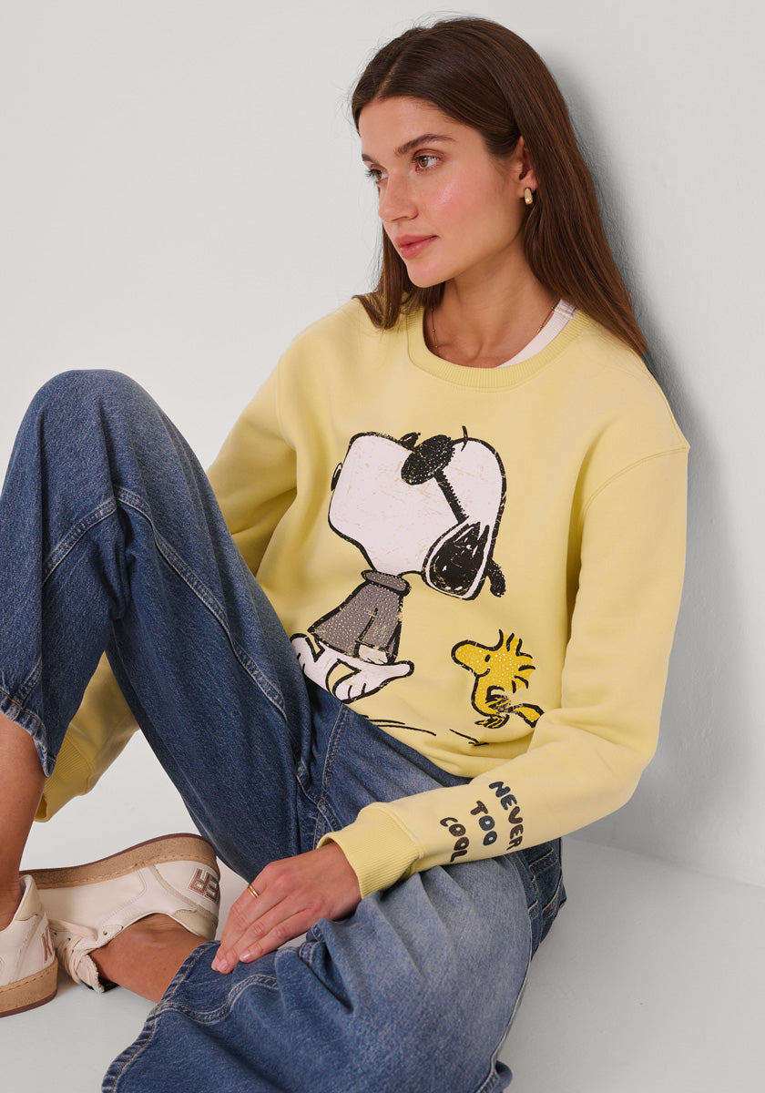 Eine Frau mit langen braunen Haaren sitzt an einer weißen Wand und trägt ein gelbes Princess Goes Hollywood Snoopy Sweatshirt mit der Aufschrift "NEVER TOO COOL" auf dem Ärmel, blaue Jeans und weiße Turnschuhe. Sie blickt nachdenklich zur Seite.