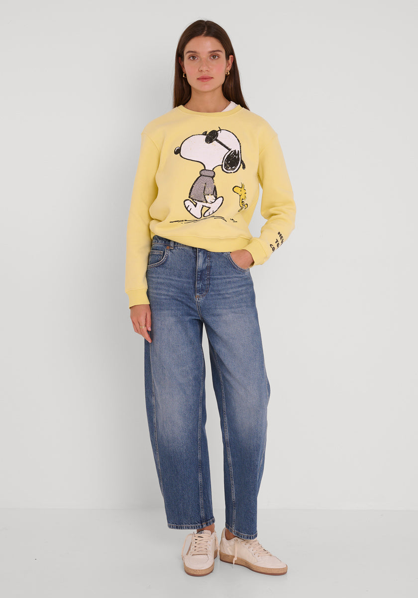 Eine Frau mit langen braunen Haaren und einem neutralen Gesichtsausdruck trägt ein hellgelbes Princess Goes Hollywood Snoopy Baumwolle Sweatshirt mit Woodstock, blaue Jeans mit hoher Taille und beigefarbene Turnschuhe. Sie steht vor einer schlichten weißen Wand und hat eine Hand in der Tasche.